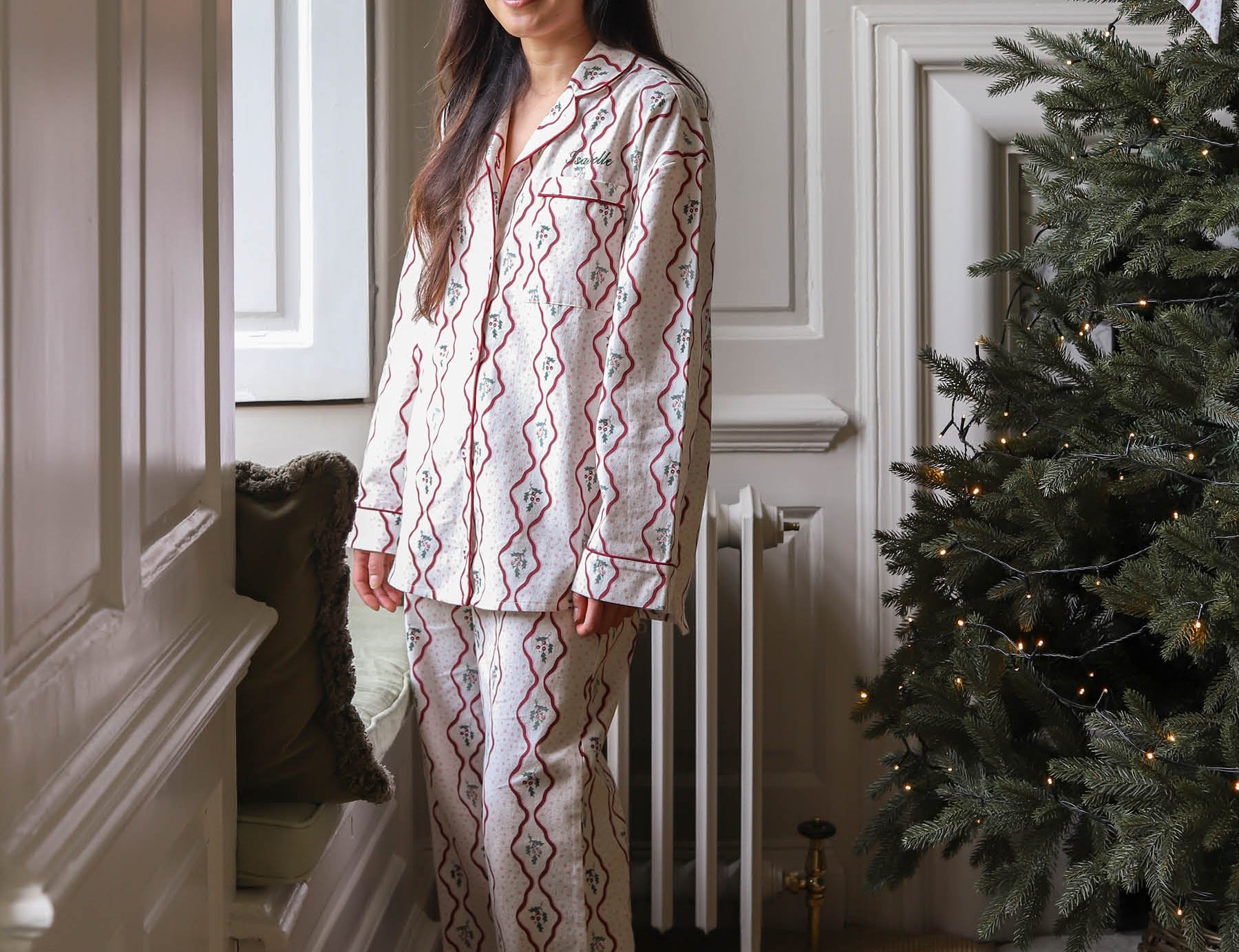 A woman wearing a two-piece pajama set with a red and blue geometric diamond pattern on a cream background, standing in a decorated room next to a Christmas tree.