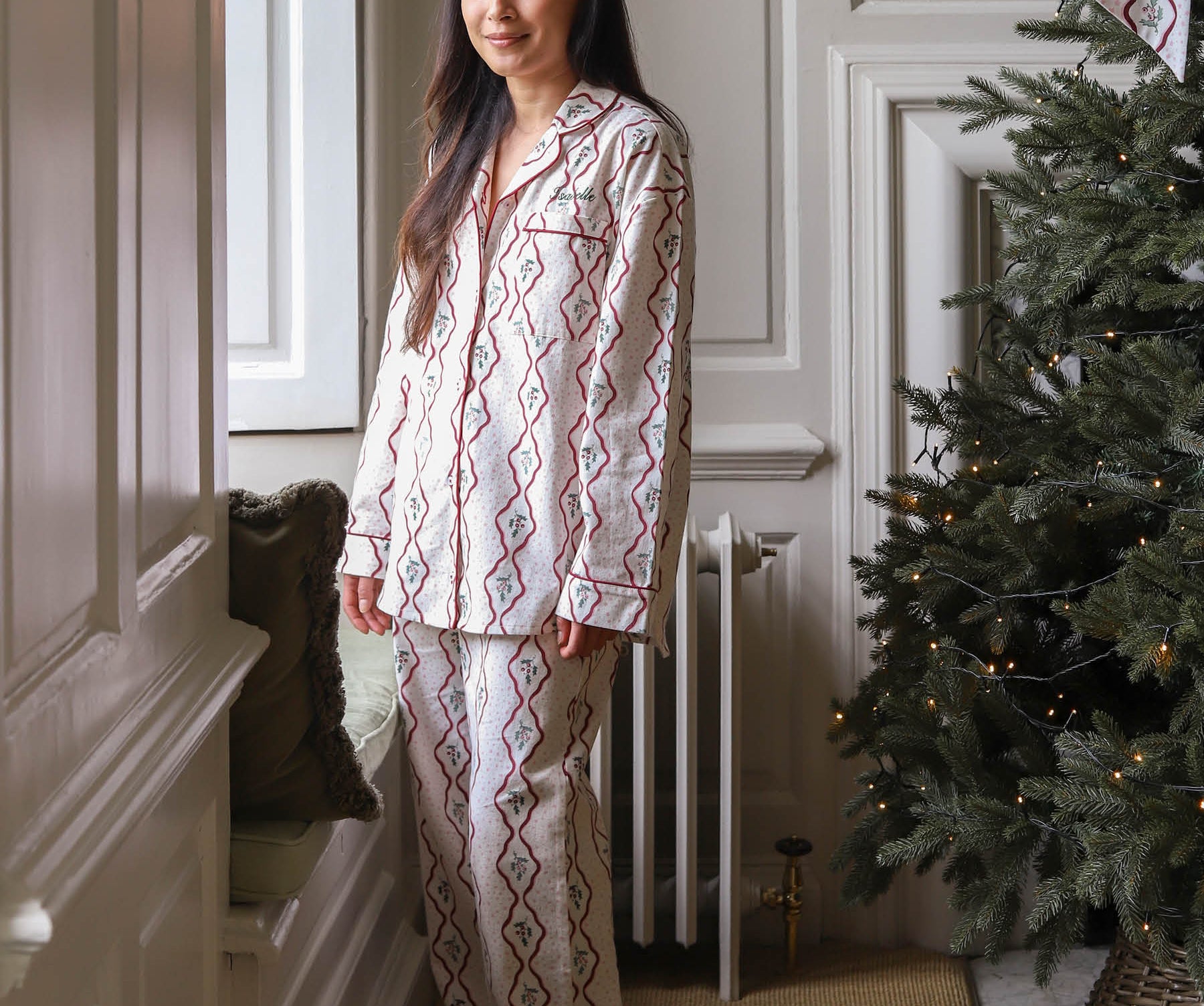 A woman wearing a two-piece pajama set with a red and blue geometric diamond pattern on a cream background, standing in a decorated room next to a Christmas tree.