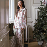 A woman wearing a two-piece pajama set with a red and blue geometric diamond pattern on a cream background, standing in a decorated room next to a Christmas tree.