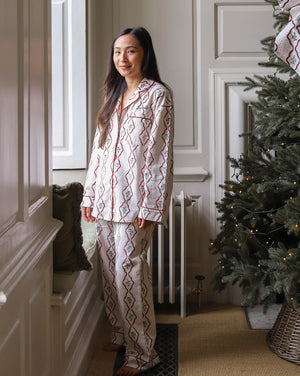 A woman wearing a two-piece pajama set with a red and blue geometric diamond pattern on a cream background, standing in a decorated room next to a Christmas tree.