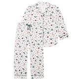 A two-piece pajama set featuring a long-sleeve button-front top and matching pants in an off-white base with a festive Christmas print of reindeer, candy canes, pine trees, and snowflakes, accented with dark green piping and a dark green drawstring.