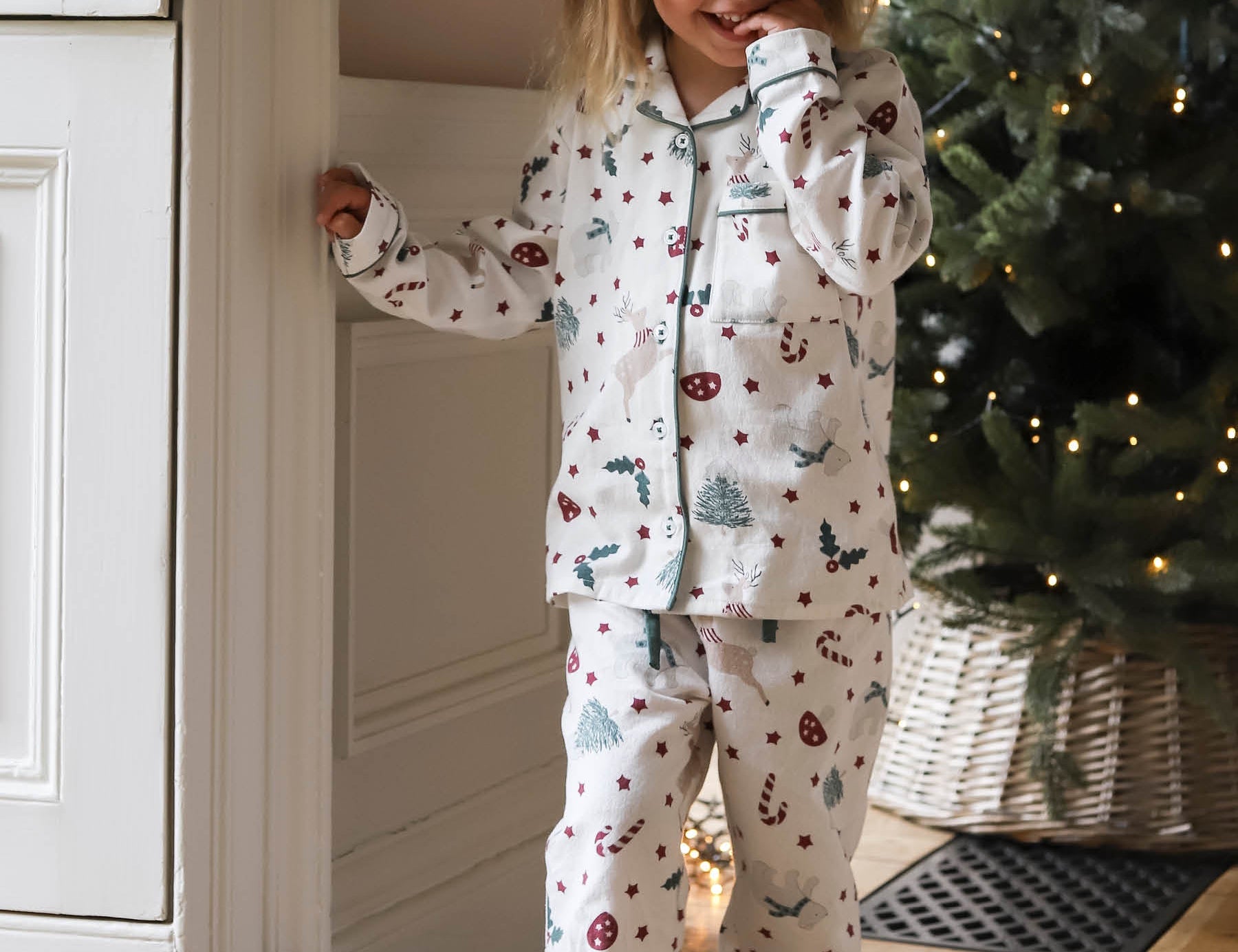 A young girl wearing a long-sleeve pajama set with a Christmas print, standing in a cozy room next to a white paneled wall and a decorated Christmas tree.