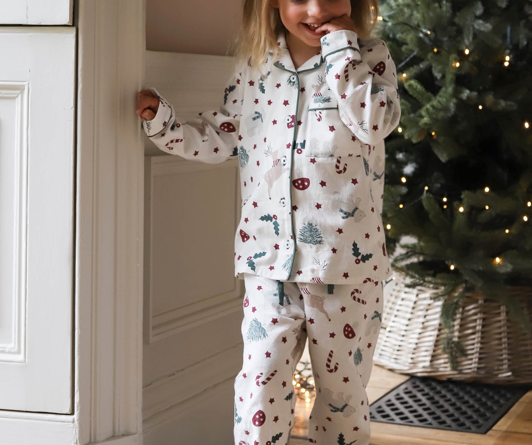 A young girl wearing a long-sleeve pajama set with a Christmas print, standing in a cozy room next to a white paneled wall and a decorated Christmas tree.