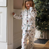 A young girl wearing a long-sleeve pajama set with a Christmas print, standing in a cozy room next to a white paneled wall and a decorated Christmas tree.