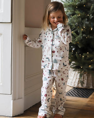 A young girl wearing a long-sleeve pajama set with a Christmas print, standing in a cozy room next to a white paneled wall and a decorated Christmas tree.