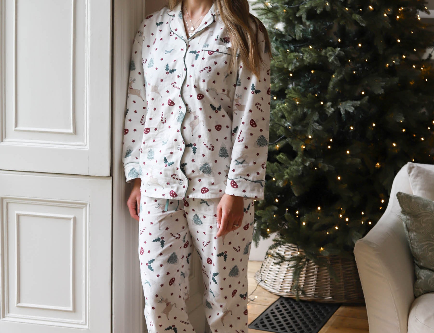 A woman stands barefoot indoors wearing a matching pajama set with a Christmas tree and berry print. The room has a white paneled door, a decorated Christmas tree with warm lights, and part of a sofa visible.