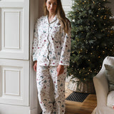 A woman stands barefoot indoors wearing a matching pajama set with a Christmas tree and berry print. The room has a white paneled door, a decorated Christmas tree with warm lights, and part of a sofa visible.
