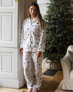 A woman stands barefoot indoors wearing a matching pajama set with a Christmas tree and berry print. The room has a white paneled door, a decorated Christmas tree with warm lights, and part of a sofa visible.