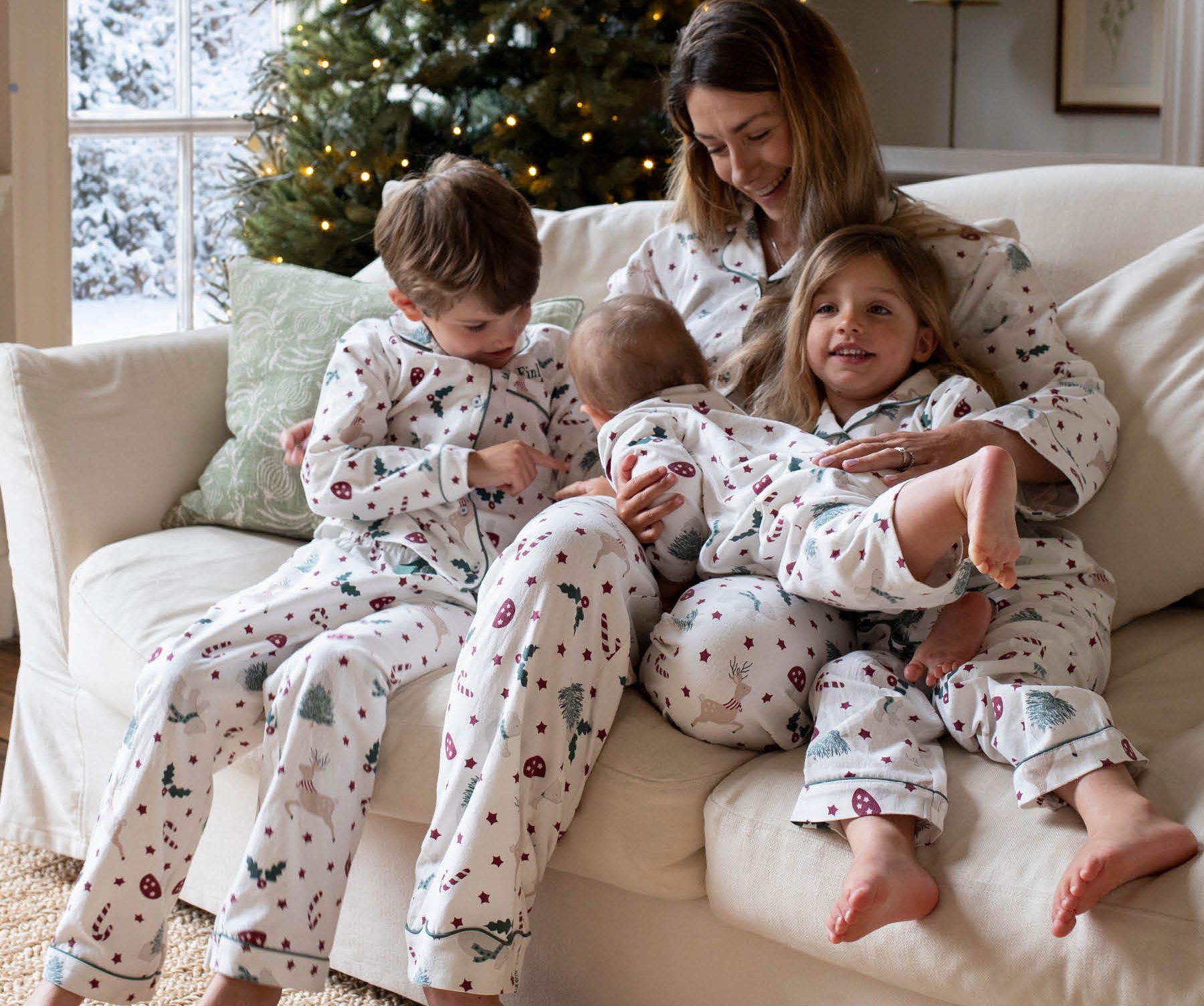 A family of four wearing matching holiday-themed pajamas sits together on a cream-colored sofa in a cozy living room with a decorated Christmas tree in the background.