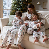 A family of four wearing matching holiday-themed pajamas sits together on a cream-colored sofa in a cozy living room with a decorated Christmas tree in the background.