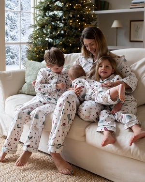 A family of four wearing matching holiday-themed pajamas sits together on a cream-colored sofa in a cozy living room with a decorated Christmas tree in the background.