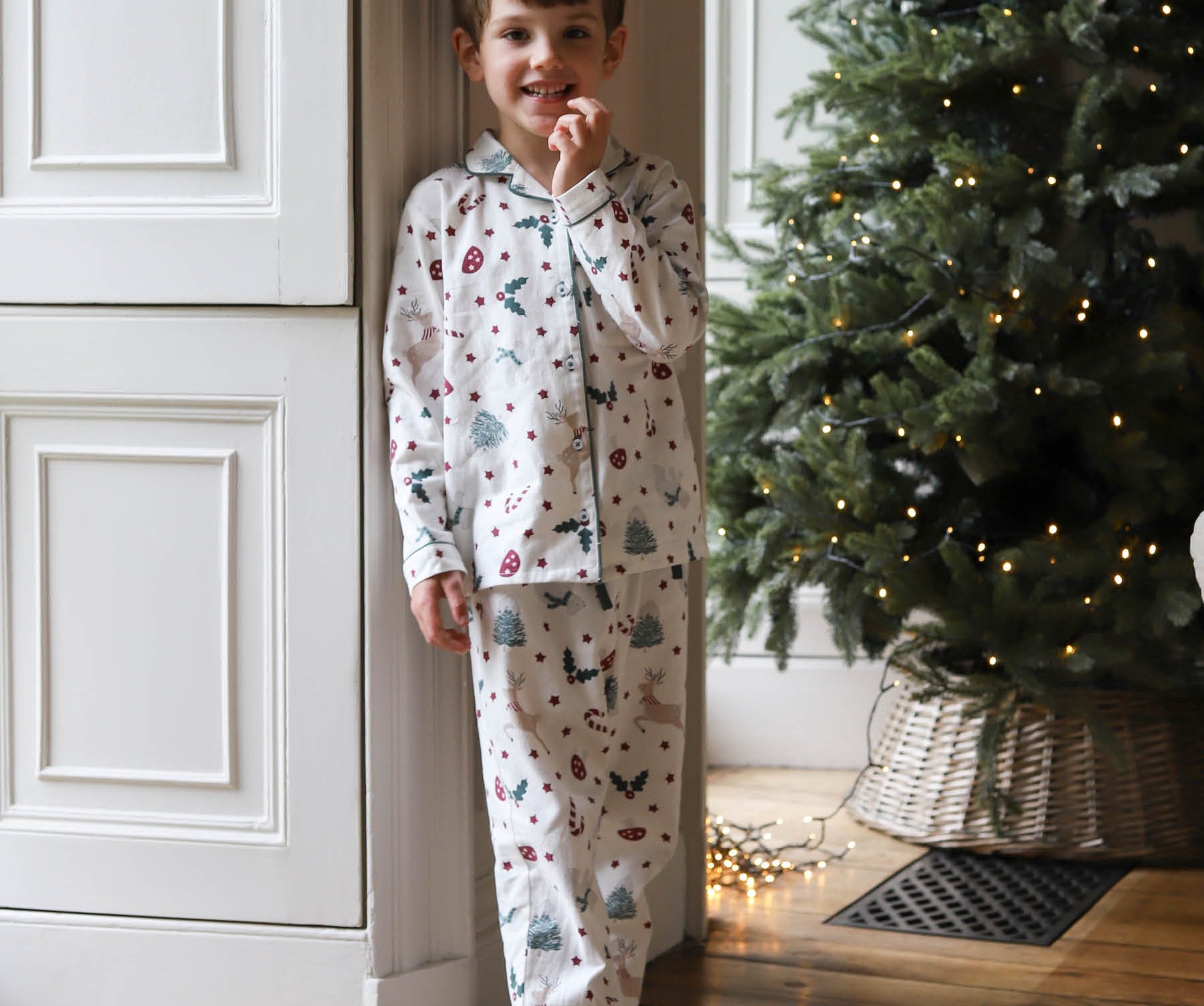A young boy wearing a white long-sleeve pajama set with a colorful Christmas tree and snowman print, standing barefoot in a doorway next to a decorated Christmas tree.