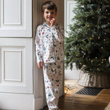 A young boy wearing a white long-sleeve pajama set with a colorful Christmas tree and snowman print, standing barefoot in a doorway next to a decorated Christmas tree.
