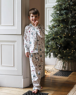 A young boy wearing a white long-sleeve pajama set with a colorful Christmas tree and snowman print, standing barefoot in a doorway next to a decorated Christmas tree.