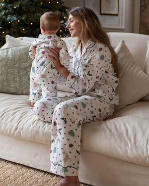 A woman and a baby wearing matching holiday-themed pajamas sit together on a cream-colored couch in a cozy living room with a Christmas tree in the background.