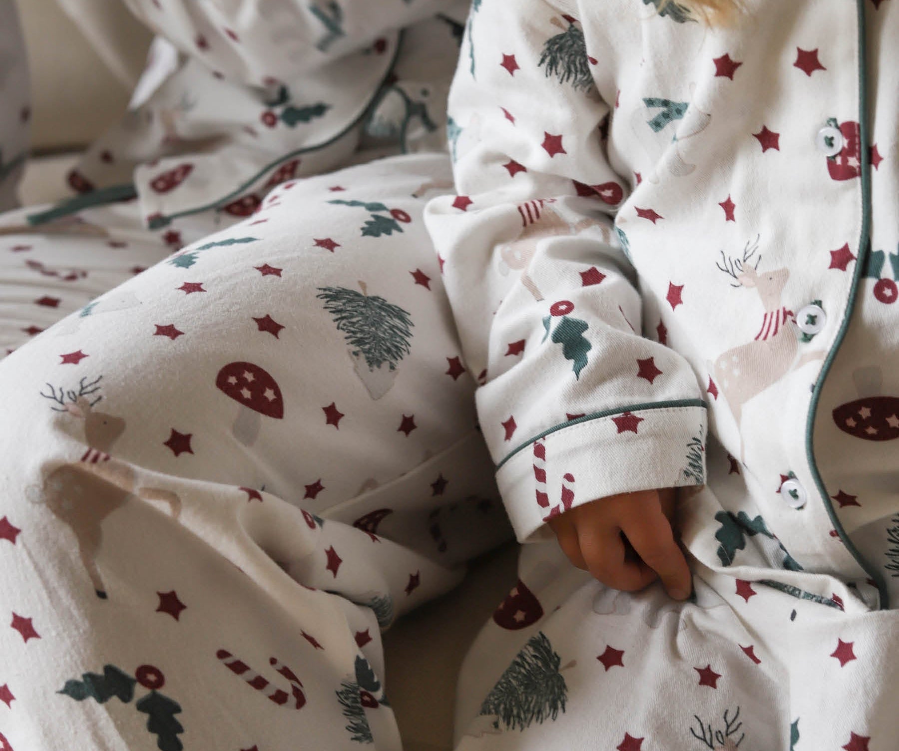 A close-up of a child wearing a cozy white Christmas-themed pajama set with red stars, green pine trees, candy canes, and reindeer motifs. The soft fabric and festive print are shown in a warm, cozy indoor setting.