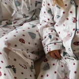 A close-up of a child wearing a cozy white Christmas-themed pajama set with red stars, green pine trees, candy canes, and reindeer motifs. The soft fabric and festive print are shown in a warm, cozy indoor setting.