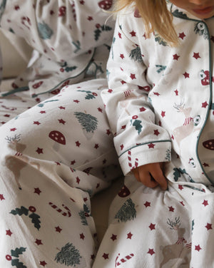 A close-up of a child wearing a cozy white Christmas-themed pajama set with red stars, green pine trees, candy canes, and reindeer motifs. The soft fabric and festive print are shown in a warm, cozy indoor setting.