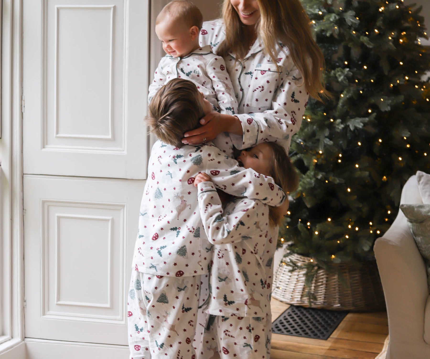 Three children and a woman wearing matching white Christmas-themed pajamas hugging in a cozy living room with a lit Christmas tree in the background.