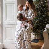 Three children and a woman wearing matching white Christmas-themed pajamas hugging in a cozy living room with a lit Christmas tree in the background.