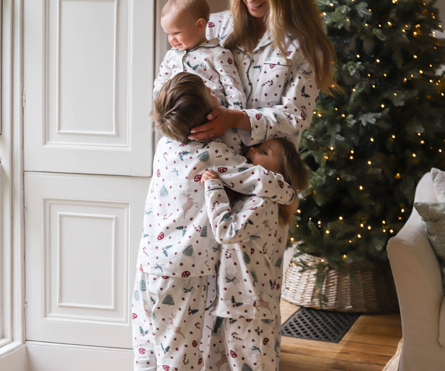 A woman and two children wearing matching Christmas-themed pajamas with a festive print, standing in a cozy room next to a decorated Christmas tree.