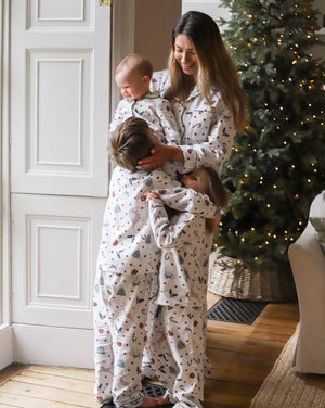 A woman and two children wearing matching Christmas-themed pajamas with a festive print, standing in a cozy room next to a decorated Christmas tree.