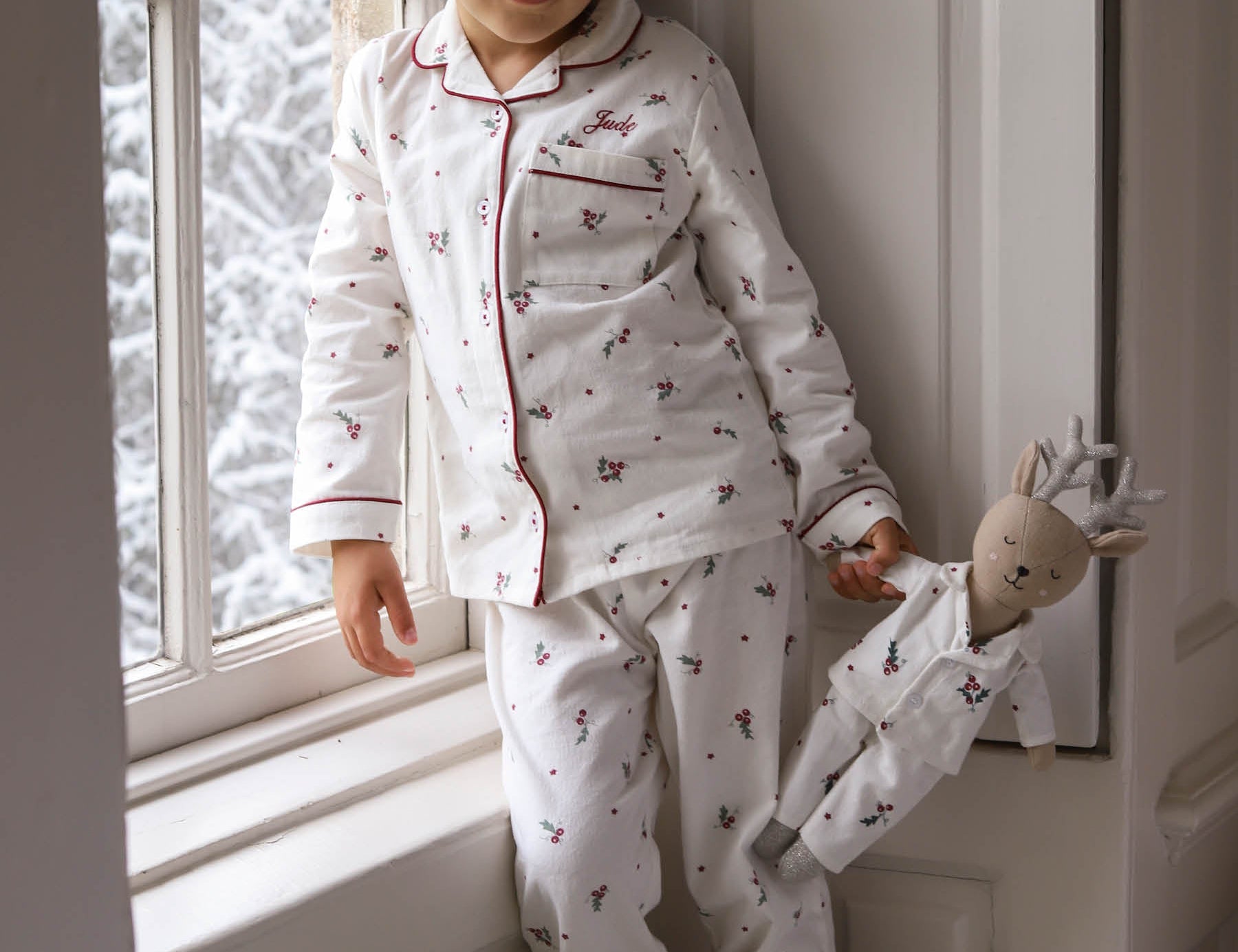 A young child wearing a white long-sleeve pajama set with a small red and dark pattern, leaning against a window frame and holding a stuffed doll.