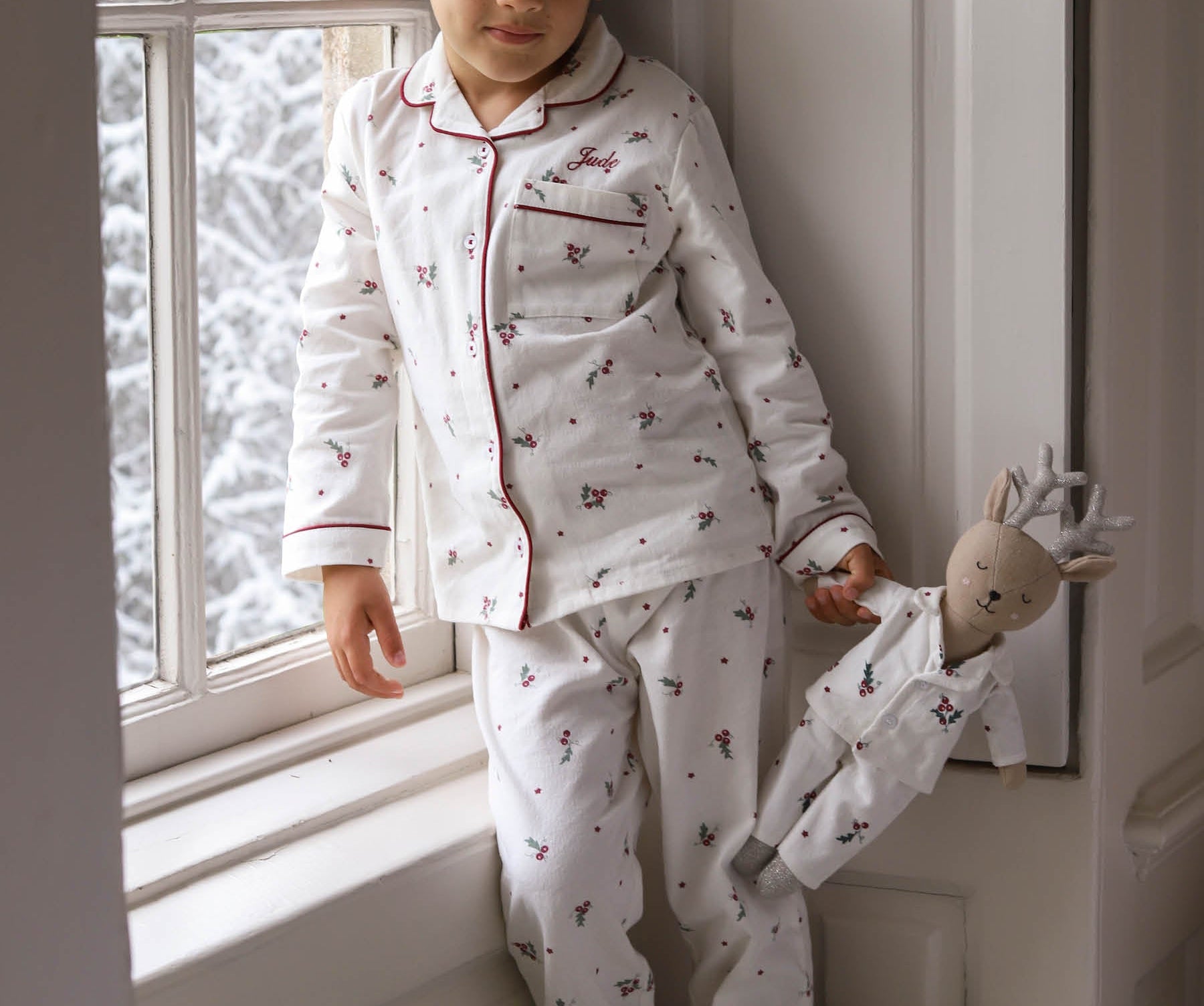 A young child wearing a white long-sleeve pajama set with a small red and dark pattern, leaning against a window frame and holding a stuffed doll.