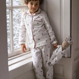 A young child wearing a white long-sleeve pajama set with a small red and dark pattern, leaning against a window frame and holding a stuffed doll.