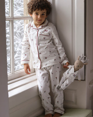 A young child wearing a white long-sleeve pajama set with a small red and dark pattern, leaning against a window frame and holding a stuffed doll.