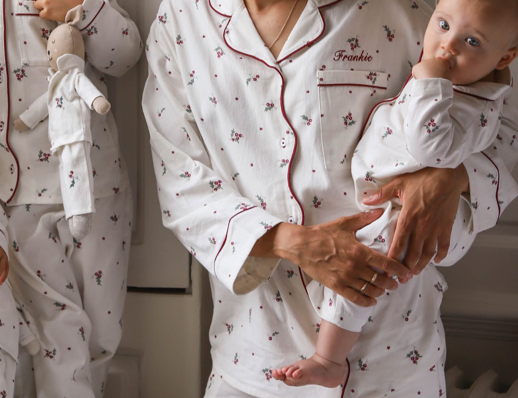 A woman and a baby wearing matching white pajamas with a small red and green floral print. The pajama set has red piping and the word 'Freddie' is embroidered on the chest pocket.