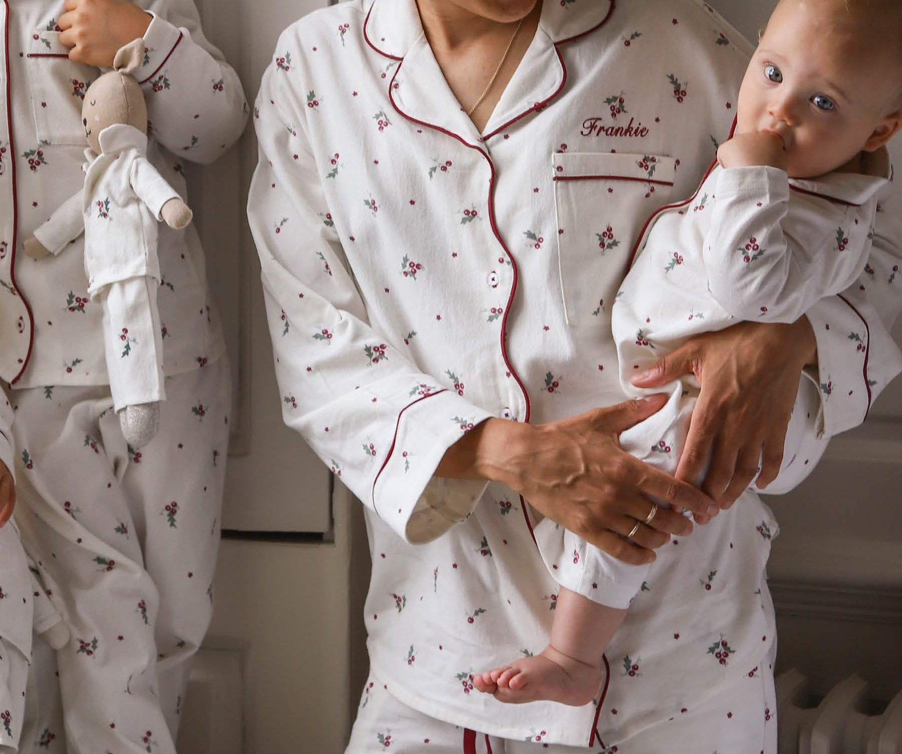 A woman and a baby wearing matching white pajamas with a small red and green floral print. The pajama set has red piping and the word 'Freddie' is embroidered on the chest pocket.
