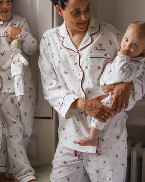 A woman and a baby wearing matching white pajamas with a small red and green floral print. The pajama set has red piping and the word 'Freddie' is embroidered on the chest pocket.