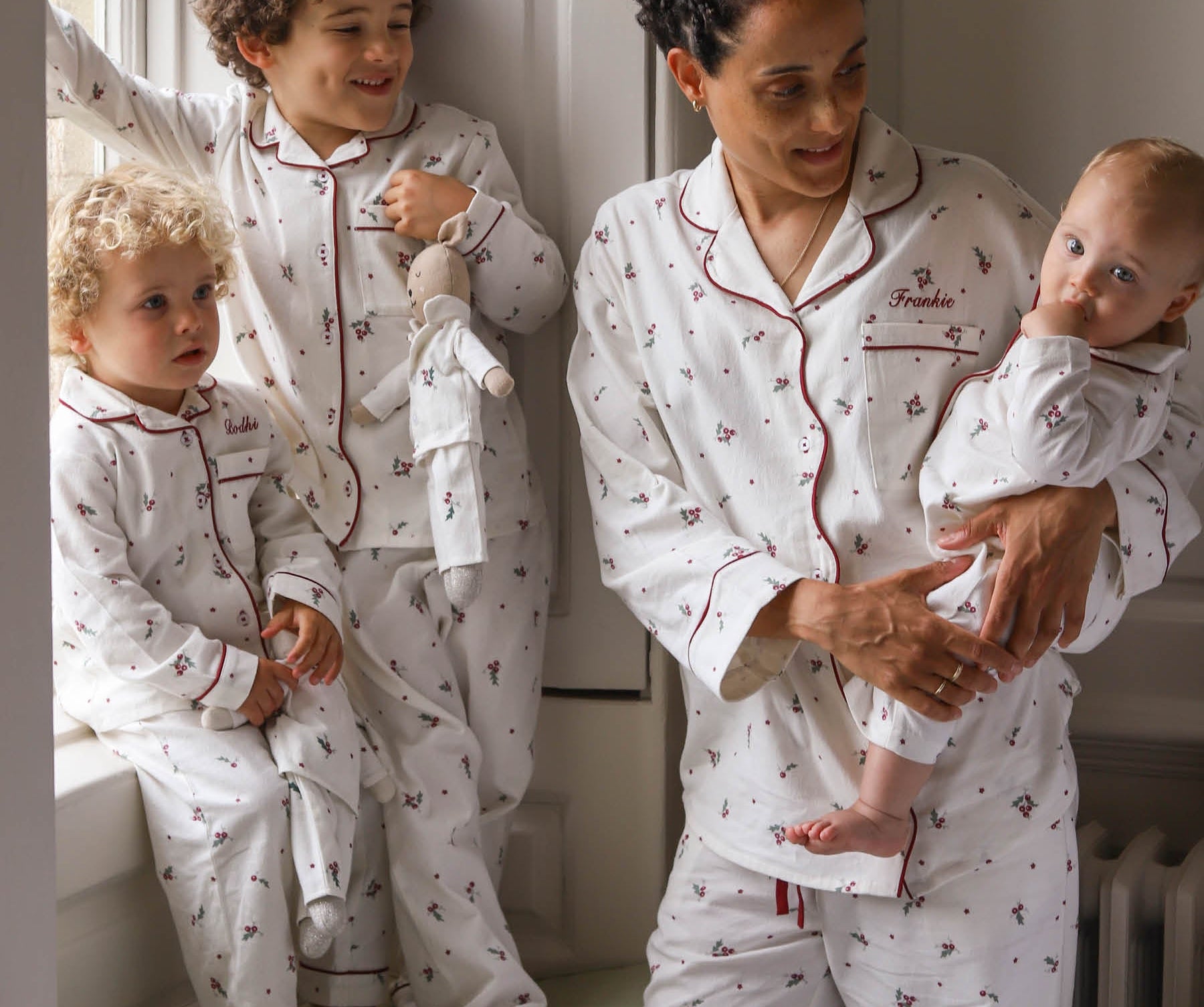 A family of four wearing matching white long-sleeve pajamas with a small red anchor pattern and red piping. The set includes a matching adult pajama set worn by a mother holding a baby, a toddler standing by a window, and a baby sitting on a window sill.