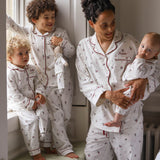A family of four wearing matching white long-sleeve pajamas with a small red anchor pattern and red piping. The set includes a matching adult pajama set worn by a mother holding a baby, a toddler standing by a window, and a baby sitting on a window sill.