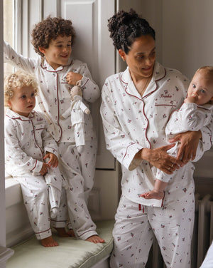 A family of four wearing matching white long-sleeve pajamas with a small red anchor pattern and red piping. The set includes a matching adult pajama set worn by a mother holding a baby, a toddler standing by a window, and a baby sitting on a window sill.