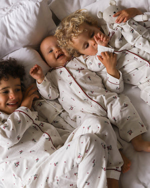 Three children lying on a bed wearing matching white long-sleeve pajamas with a small red and green floral print and dark red piping. One child is holding a white teddy bear.