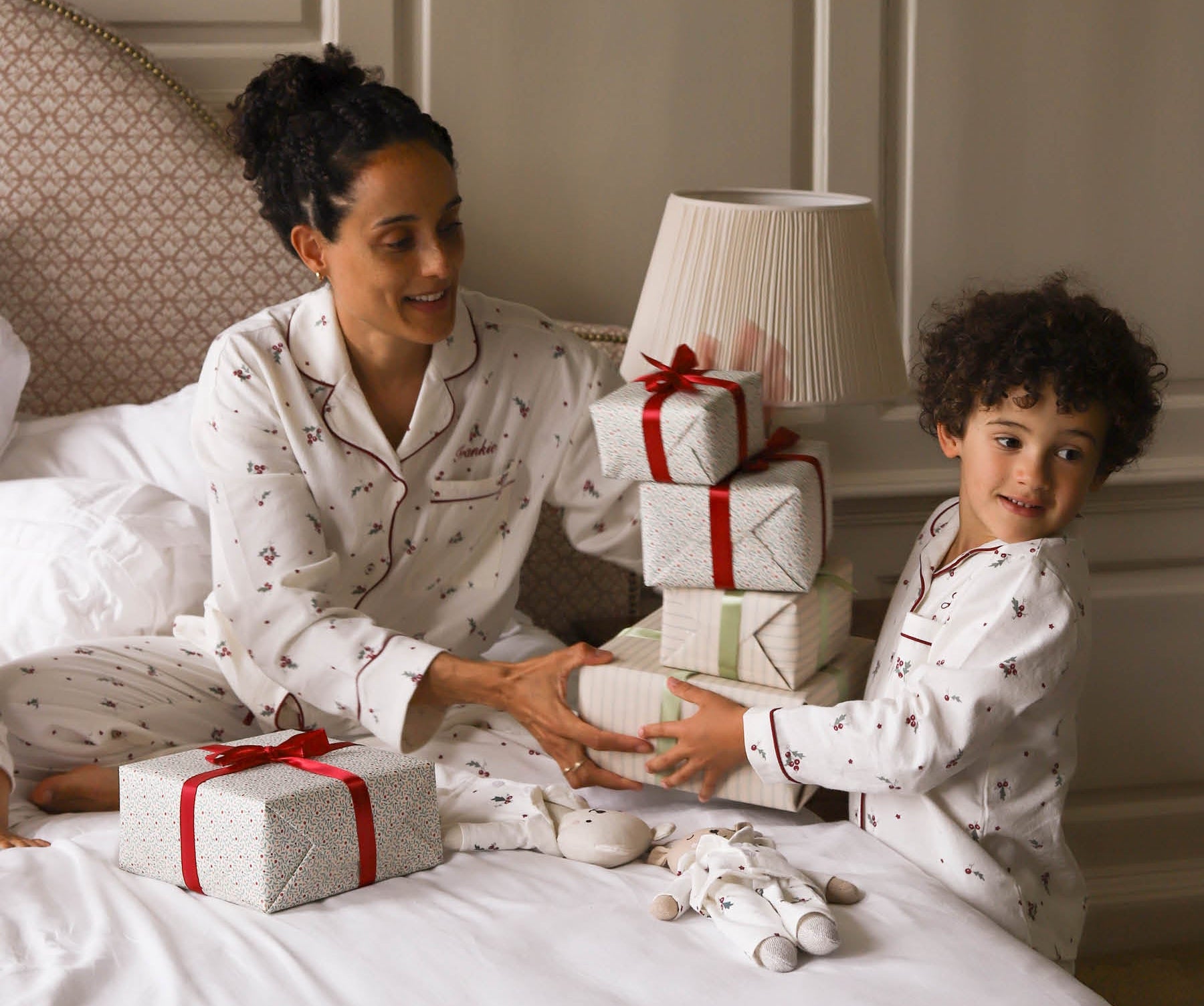 A woman and a young boy wearing matching white Christmas-themed pajamas with small red and dark motifs, sitting on a bed with wrapped gift boxes and a stack of presents.