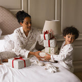A woman and a young boy wearing matching white Christmas-themed pajamas with small red and dark motifs, sitting on a bed with wrapped gift boxes and a stack of presents.