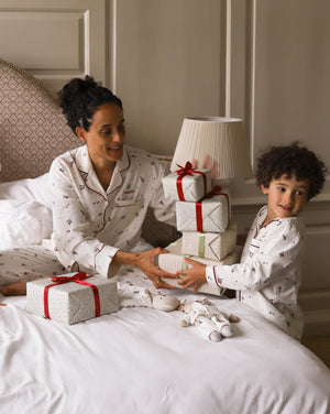 A woman and a young boy wearing matching white Christmas-themed pajamas with small red and dark motifs, sitting on a bed with wrapped gift boxes and a stack of presents.