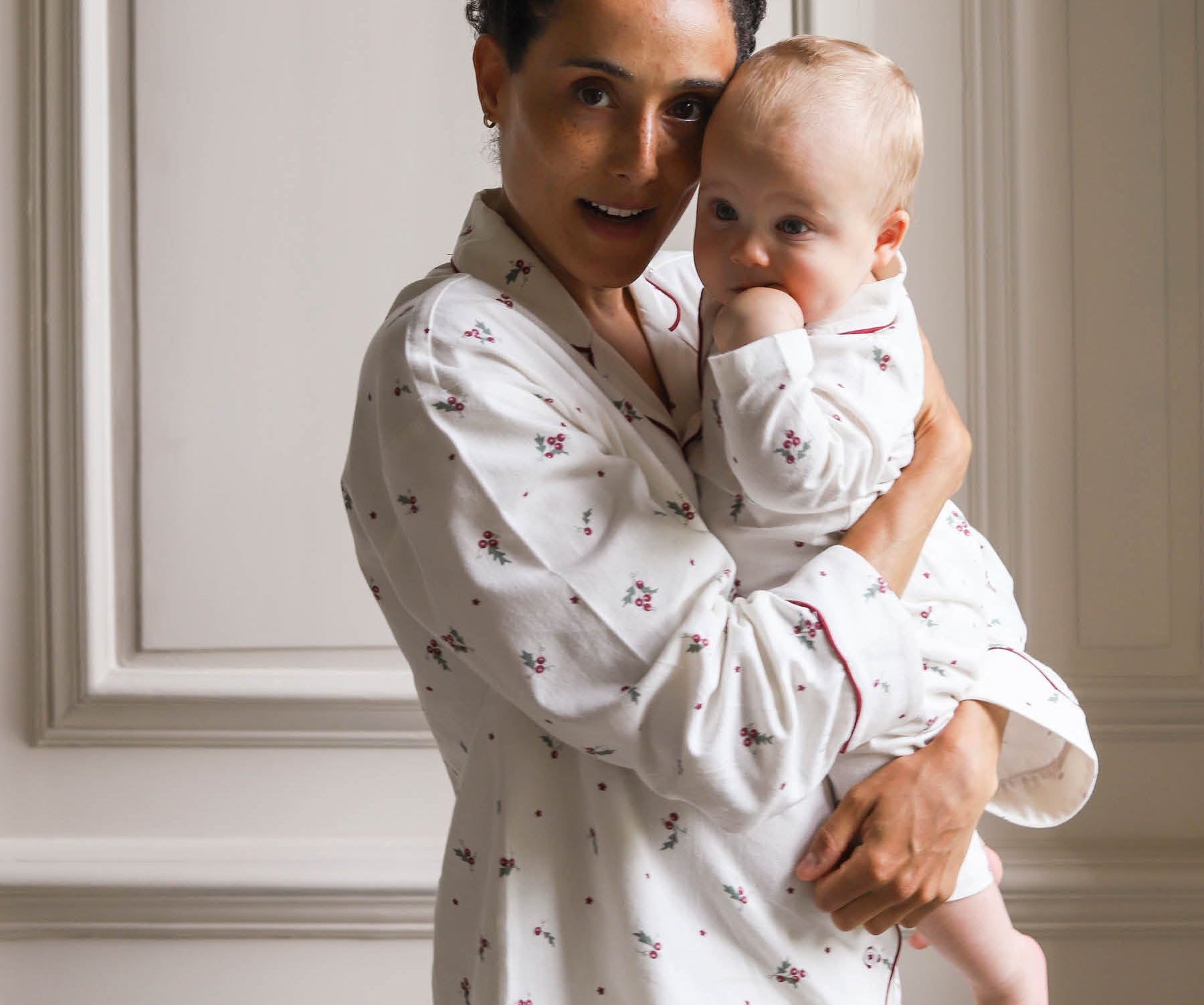 A woman holding a baby, both wearing matching white long-sleeve pajamas with a small red floral pattern, posed against a light beige paneled wall.