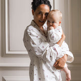 A woman holding a baby, both wearing matching white long-sleeve pajamas with a small red floral pattern, posed against a light beige paneled wall.