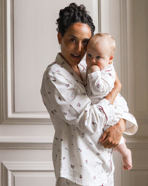 A woman holding a baby, both wearing matching white long-sleeve pajamas with a small red floral pattern, posed against a light beige paneled wall.