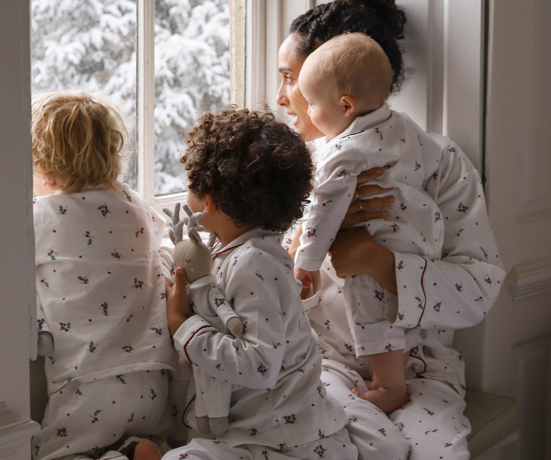 A family of four wearing matching white pajamas with a small dark pattern, sitting by a window with snow-covered trees outside. The adults and children are shown in a cozy indoor setting.