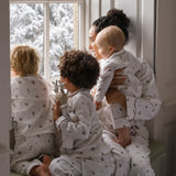 A family of four wearing matching white pajamas with a small dark pattern, sitting by a window with snow-covered trees outside. The adults and children are shown in a cozy indoor setting.