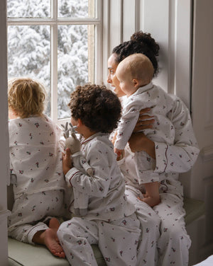 A family of four wearing matching white pajamas with a small dark pattern, sitting by a window with snow-covered trees outside. The adults and children are shown in a cozy indoor setting.