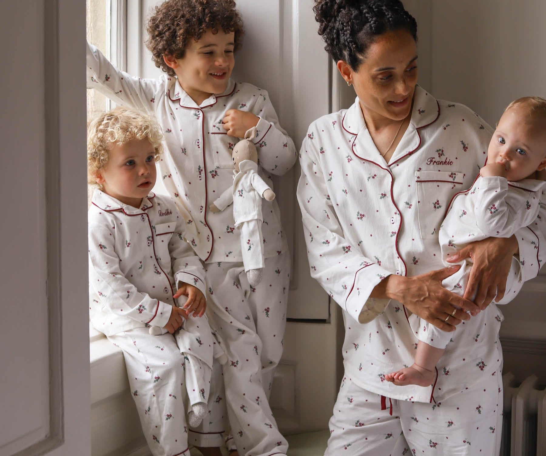 A family wearing matching white patterned pajamas, including a woman holding a baby, a standing child, and a toddler by a window.