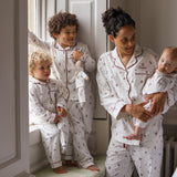 A family wearing matching white patterned pajamas, including a woman holding a baby, a standing child, and a toddler by a window.
