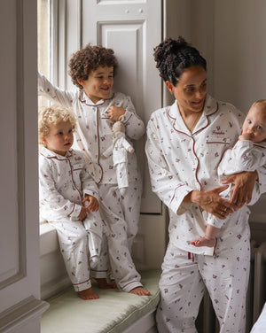 A family wearing matching white patterned pajamas, including a woman holding a baby, a standing child, and a toddler by a window.