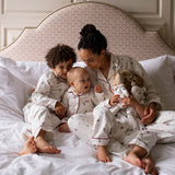 A family of four wearing matching white patterned pajamas sits together on a bed with white linens and a patterned upholstered headboard. The woman holds a doll, and the children look toward the camera.